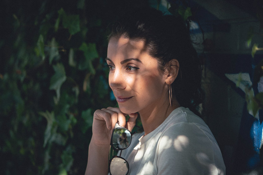 A model stands in the shade of a tree with dappled light falling across her face