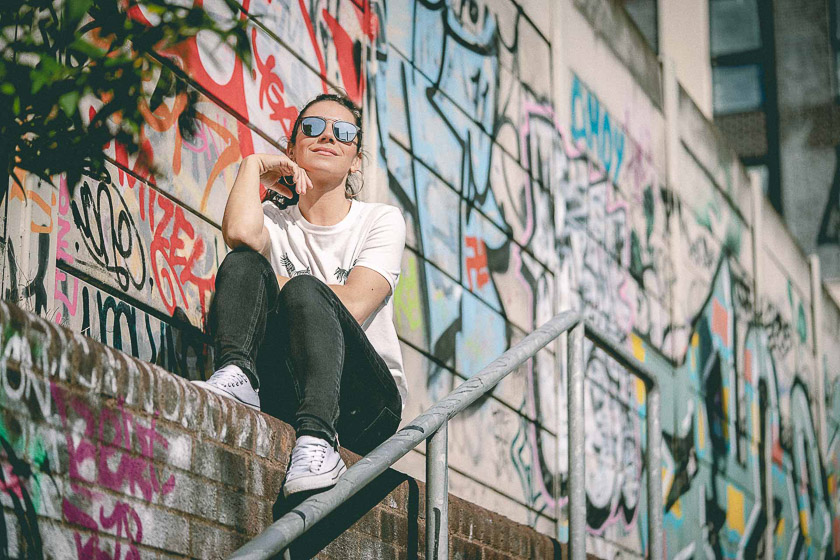 A model wearing sunglasses sits on a wall covered in colourful graffiti