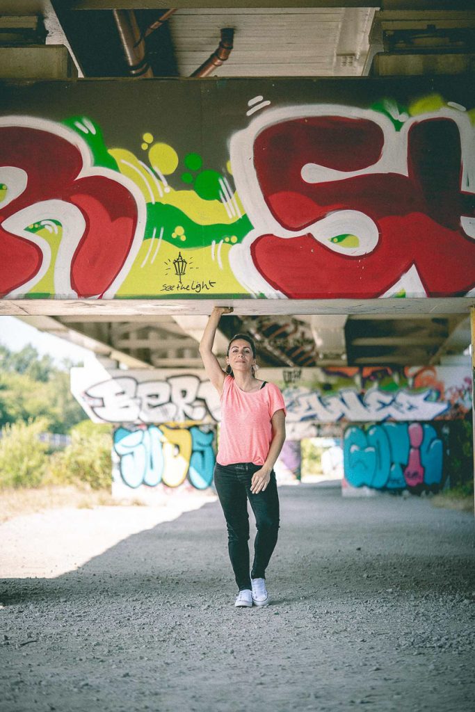 A model stands under a tram bridge that is covered in colourful graffiti