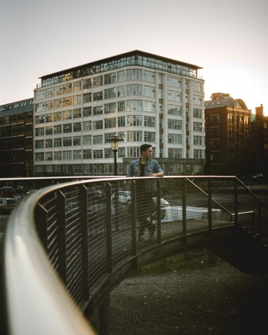 A man stands on a curved bridge in front of a white art-deco apartment block.