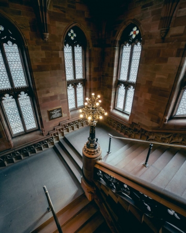 A grand stone staircase turns around an elegant light, flanked by large gothic windows