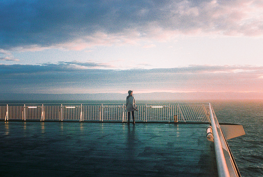 A woman stands on the deck of a ferry at sunset, looking out to sea